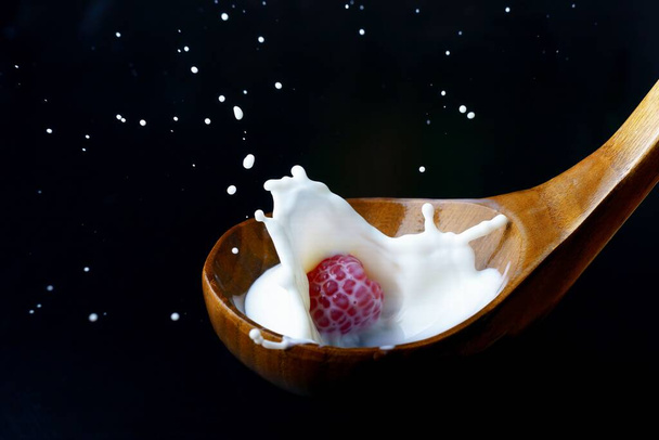 fresh raspberry splashing milk on a wooden spoon, splash effect with black background - Photo, image
