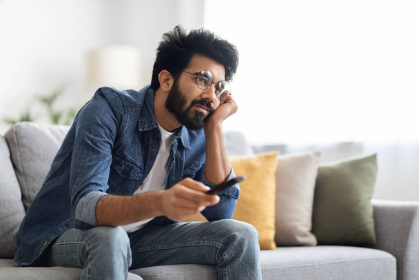 Виставка нудьги. Portrait Of Upset Young Indian Guy Watching TV At Home, Bored Eastern Man Holding Remote Controller And Switching Channels On Television, Sitting On Couch In Living Room, Copy Space - Фото, зображення