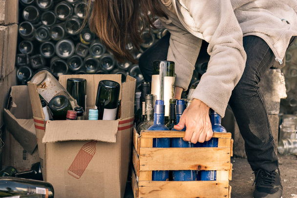 Unrecognizable woman hand holding wooden box with glass materials for recycling, in the background out of focus are many stacked glass bottles. - Фото, зображення