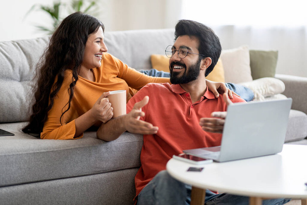 Cheerful Indian Couple Relaxing Together With Laptop And Coffee In Living Room, Happy Young Eastern Spouses Resting At Home, Having Hot Drinks, Using Computer And Chatting, Closeup Shot - Фото, изображение