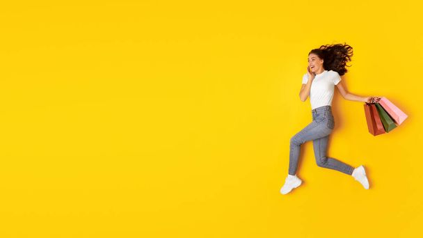 Wow Shopping Offer. Excited Woman Carrying Colorful Paper Shopper Bags, Posing Running In Mid Air Over Yellow Studio Background. Shopaholic Lady Looking At Free Space For Text. Panorama - Foto, Imagen