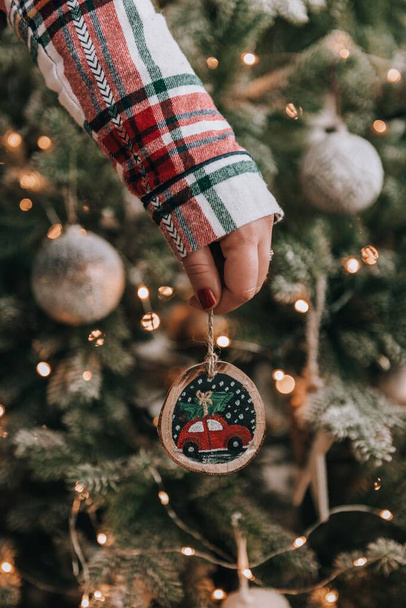 Close-up photo of woman wearing plaid pyjamas, holding diy christmas ornament in front of christmas tree. - Foto, imagen