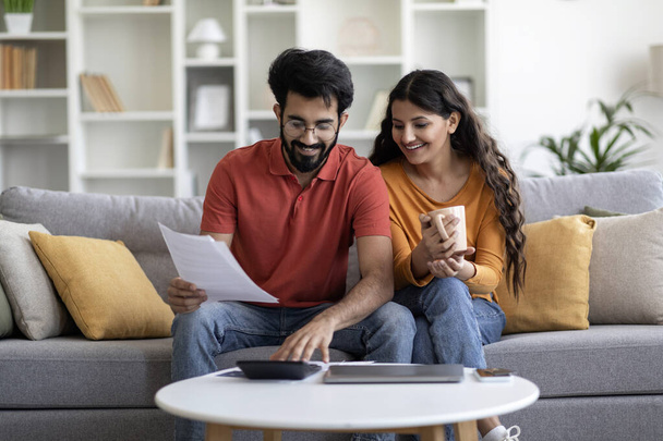 Indian Married Couple Planning Budget Together, Reading Papers And Calculating Spends While Sitting On Couch In Living Room, Young Eastern Spouses Checking Documents And Accounting Taxes, Free Space - Foto, Imagen