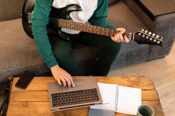 Nierozpoznawalny młody facet gra na gitarze elektrycznej i za pomocą przycisku naciśnięcia laptopa w domu, przycięty strzał. Muzyk uczący się grać na instrumencie muzycznym online za pośrednictwem komputera. Muzyka i technologia - Zdjęcie, obraz