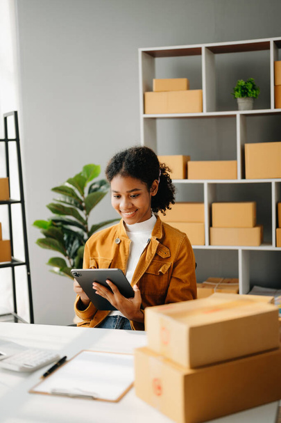 Startup small business SME, Entrepreneur owner African woman using  tablet taking receive and checking online purchase shopping order to prepare pack product boxes. in home office - Foto, Imagem