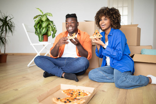 Joyeux conjoints noirs mangeant de la pizza et regardant la télévision après avoir déménagé dans une nouvelle maison, heureux jeune couple afro-américain famille mangeant des collations savoureuses et se détendre sur le sol parmi les boîtes en carton - Photo, image