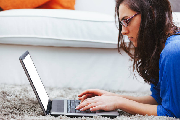 Eine junge Frau auf dem weichen und bequemen Teppich ihres Hauses benutzt einen Laptop. Arbeitet sie, studiert sie oder hat sie Spaß? Dank Wifi und Breitband, die das Internet schnell erreichen, hat sie - Foto, Bild