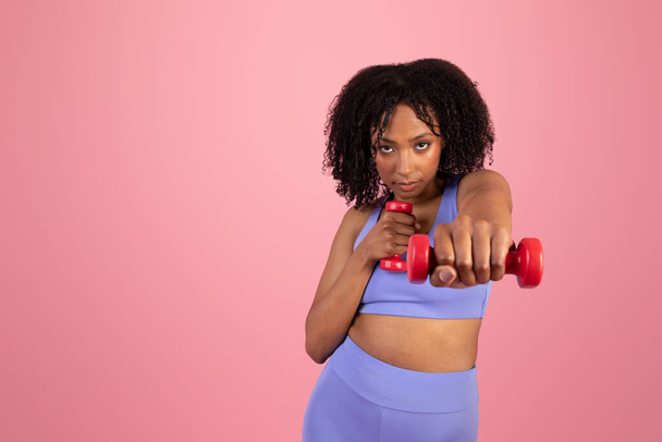 Komoly magabiztos afro-amerikai hölgy a sportruházatban, kézgyakorlatozik súlyzókkal, élvezi a sportot egyedül a rózsaszín stúdió hátterében. Alkalmas életmód, testápolás, edzés és vitalitás - Fotó, kép