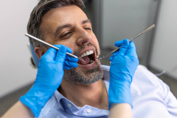 Πάνω από τον ώμο ενός οδοντιάτρου που εξετάζει τα δόντια ενός ασθενούς στην οδοντιατρική κλινική. Εξετάζει τα δόντια του ένας νεαρός οδοντίατρος.. - Φωτογραφία, εικόνα