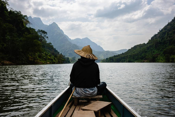 The Asian boy with a straw hat on the riverboat  on the Nam Ou River in the Mountains of Nong Khiaw, Laos, backside view - Zdjęcie, obraz