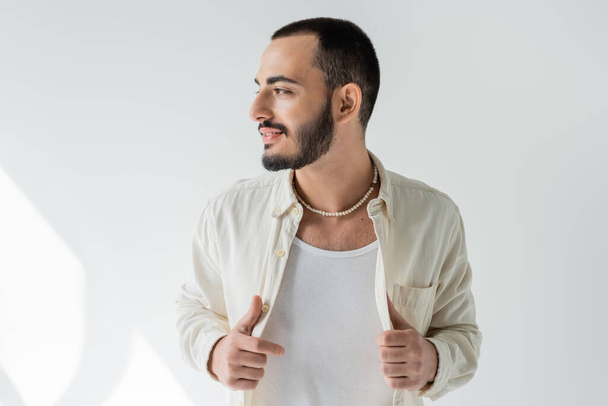 Smiling and bearded gay man in casual clothes and pearl necklace touching shirt and looking away while posing on grey background with sunlight  - Фото, зображення