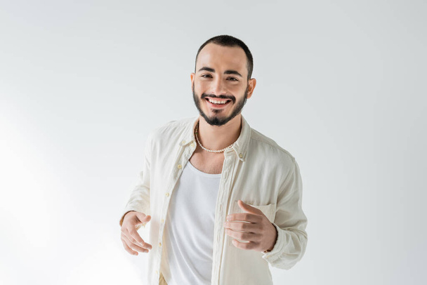 Cheerful and bearded young homosexual man in beige shirt and pearl necklace looking at camera while posing on grey background - Photo, Image