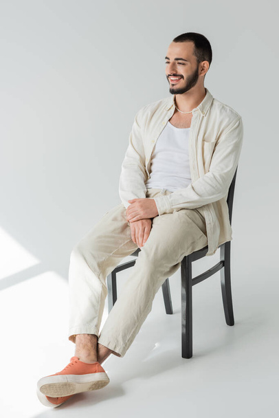 Homem homossexual despreocupado em colar de pérolas e roupas casuais feitas de tecidos naturais desviando o olhar enquanto sentado na cadeira em fundo cinza com luz solar  - Foto, Imagem
