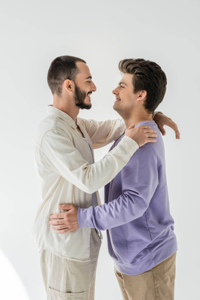 Side view of young and cheerful homosexual couple in casual clothes hugging and looking at each other while standing on grey background - Fotoğraf, Görsel
