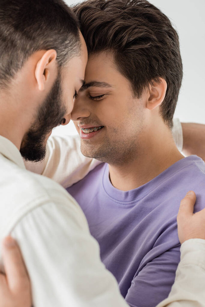 Portrait of positive brunette gay man in braces hugging bearded partner in shirt while closing eyes and standing nose to nose isolated on grey  - Foto, Bild