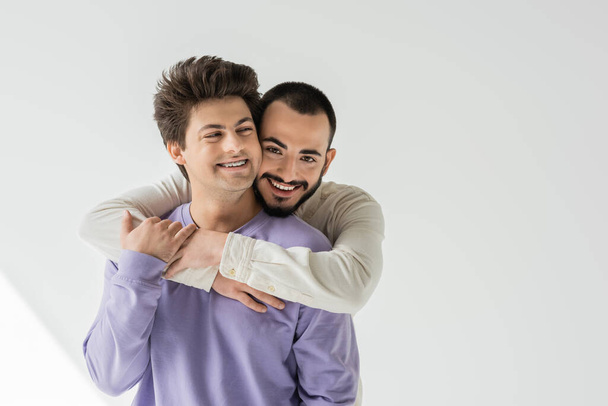 Allegro e barbuto uomo omosessuale che abbraccia il giovane partner bruna in bretelle e felpa viola mentre guarda la fotocamera su sfondo grigio con la luce del sole  - Foto, immagini
