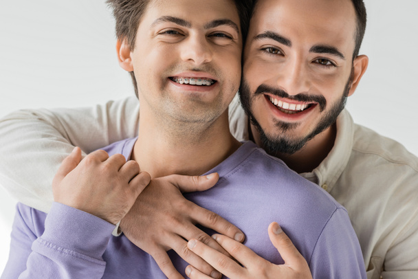 Portrait of young and cheerful same sex couple in casual clothes hugging and touching hands while looking at camera isolated on grey  - Fotoğraf, Görsel