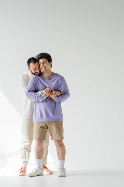 Smiling and bearded homosexual man in casual clothes holding hands of young boyfriend with closed eyes while standing on grey background with sunlight  - Photo, image