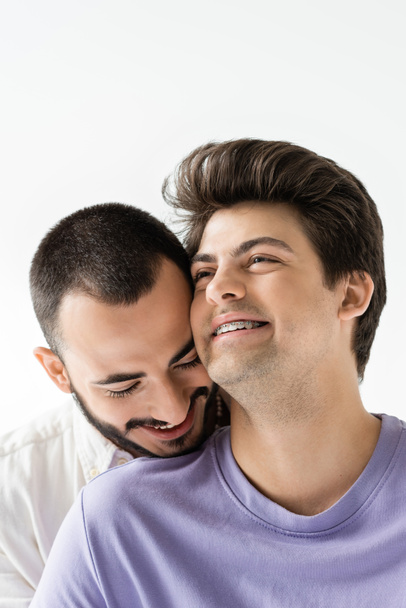 Portrait of brunette and positive gay man with braces on teeth looking away while standing near bearded boyfriend isolated on grey  - Foto, Imagem
