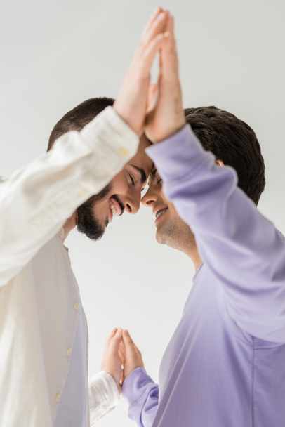 Низький кут зору молодої і усміхненої гомосексуальної пари в повсякденному одязі закриває очі, тримаючись за руки і стоячи разом ізольовано на сірому
  - Фото, зображення