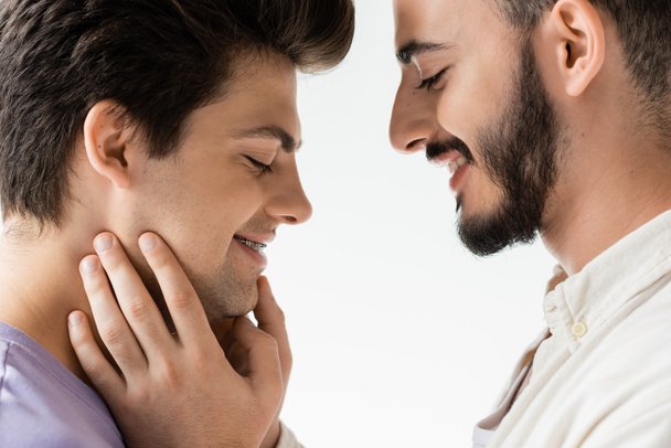 Боковой вид позитивного бородатого гея в повседневной рубашке трогательное лицо молодого партнера с брекетами на зубах, стоя вместе изолированы на сером  - Фото, изображение