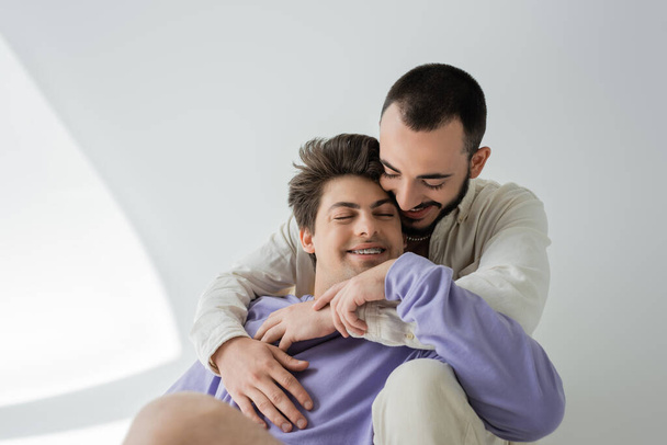 Joven morena gay hombre abrazando despreocupado novio con los ojos cerrados, en frenillos y en sudadera púrpura mientras se sientan juntos sobre fondo gris con la luz del sol - Foto, imagen
