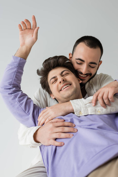 Positive and bearded homosexual man in casual clothes hugging brunette partner with closed eyes and braces while sitting together isolated on grey  - Fotoğraf, Görsel