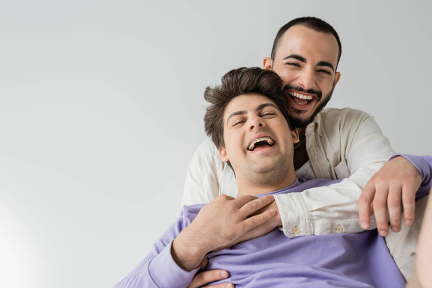 Der bärtige homosexuelle Mann im Hemd umarmt und berührt die Hand seines lachenden Freundes in Hosenträgern und lila Sweatshirt, während er isoliert auf grau sitzt  - Foto, Bild