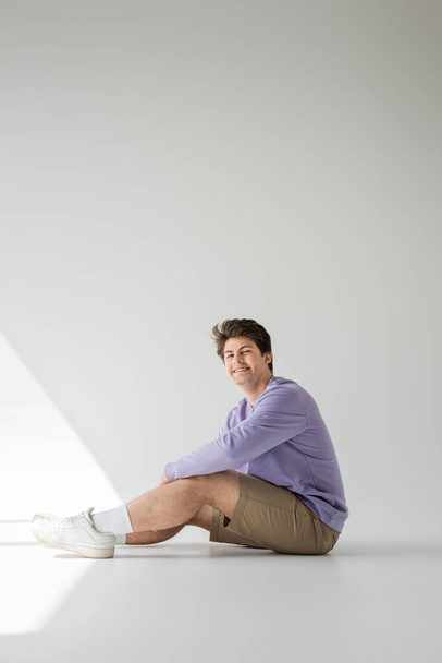 Pełna długość uśmiechniętego homoseksualisty w aparacie fotograficznym, beżowych spodenkach i fioletowej bluzie, siedzącego na szarym tle ze światłem słonecznym  - Zdjęcie, obraz