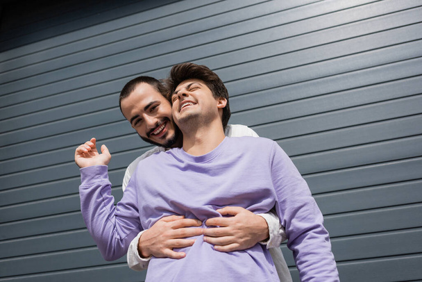 Low angle view of positive and bearded gay man hugging brunette boyfriend in sweatshirt and braces while looking at camera near building on urban street  - Foto, afbeelding