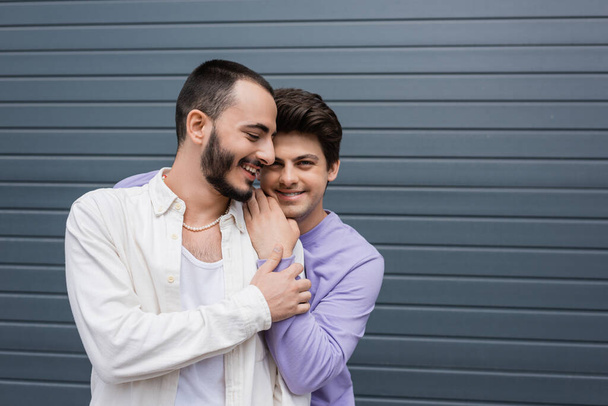 Smiling brunette gay man in sweatshirt and braces hugging partner and looking at camera while standing together near grey building outdoors on urban street  - Photo, Image