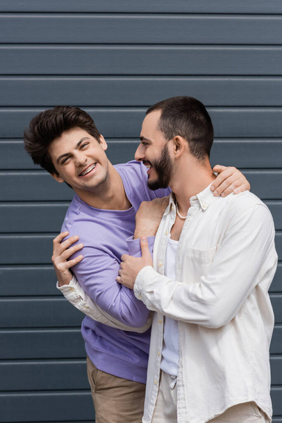 Portrait of cheerful homosexual man in purple sweatshirt and braces hugging young bearded boyfriend and looking at camera while standing near building outdoors  - Φωτογραφία, εικόνα