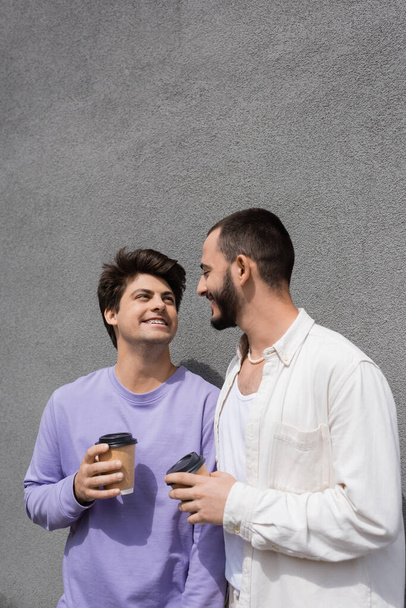 Bearded homosexual man holding coffee to go and looking at smiling boyfriend in braces while talking near building on urban street at daytime - Фото, изображение