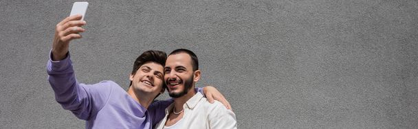 Alegre pareja homosexual en ropa casual tomando selfie en el teléfono inteligente juntos mientras están de pie cerca de la pared gris del edificio en la calle urbana durante el día, pancarta  - Foto, Imagen