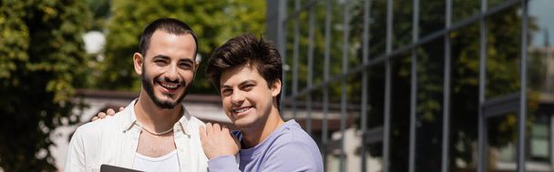Smiling homosexual man in casual clothes hugging bearded boyfriend and looking at camera together outdoors at daytime, on urban street, banner  - Photo, image