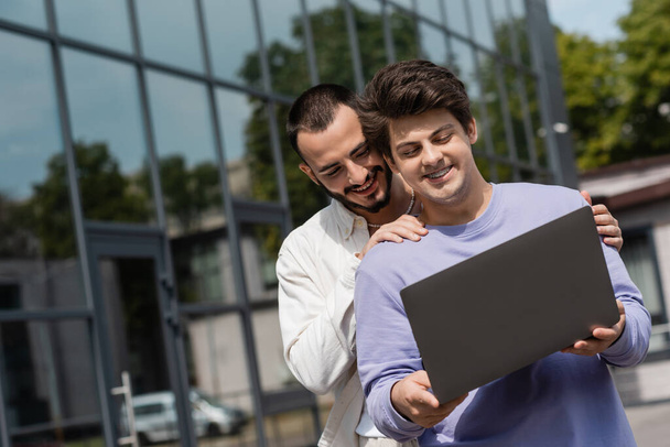 Overjoyed and bearded man hugging smiling boyfriend with braces in casual clothes using laptop while standing together near blurred building on urban street  - Foto, Bild