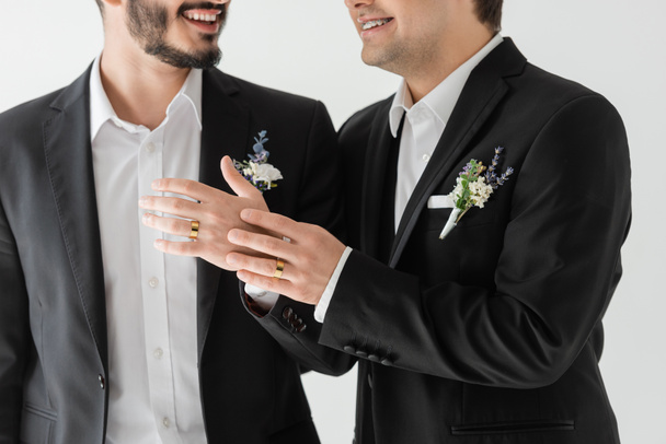 Cropped view of smiling gay groom in elegant suits with floral boutonniere touching hand of boyfriend in braces with wedding ring while standing isolated on grey   - Zdjęcie, obraz