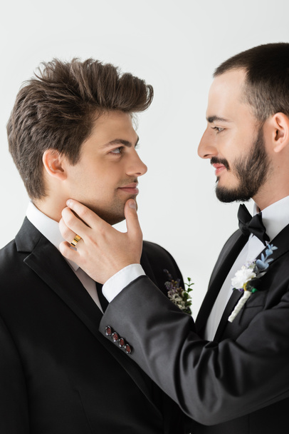 Portrait of carefree homosexual groom in formal wear with floral boutonniere touching chin of young boyfriend during wedding celebration isolated on grey   - Zdjęcie, obraz