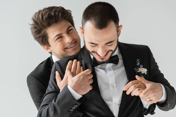 Portrait d'homme homosexuel positif avec bretelles en costume élégant et anneau d'or à la main étreignant marié barbu lors de la célébration du mariage isolé sur gris   - Photo, image