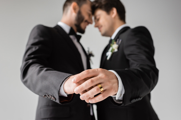 Vue latérale d'un couple de même sexe flou en tenue formelle avec des boutonnières se touchant les mains avec anneau doré pendant le mariage isolé sur gris  - Photo, image