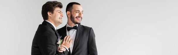 Pozitivní homosexuál v obleku s boutonniere dotýkat vousatý přítel a dívat se pryč během svatebního obřadu izolované na šedé, banner  - Fotografie, Obrázek