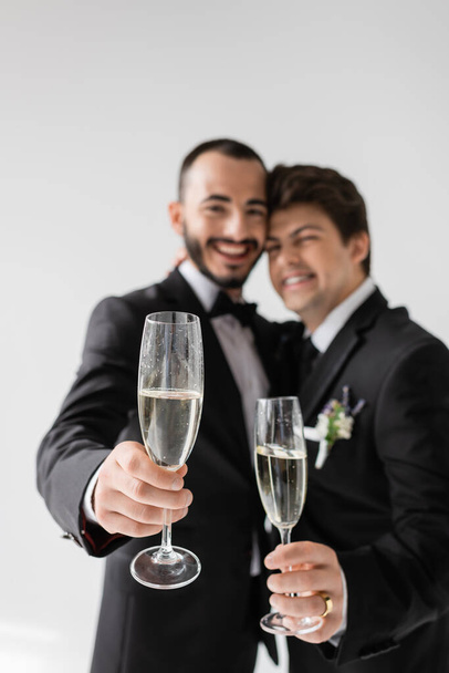 Blurred lgbt couple in elegant formal wear smiling and showing champagne glasses at camera during wedding celebration isolated on grey  - Foto, afbeelding