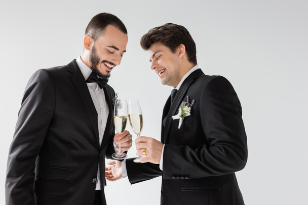 Tasasız ve sakallı homoseksüel damat, gri renkte izole edilmiş düğün töreninde diş telli, yaka çiçekli zarif erkek arkadaşıyla şampanya kadehleri kaldırıyor.  - Fotoğraf, Görsel
