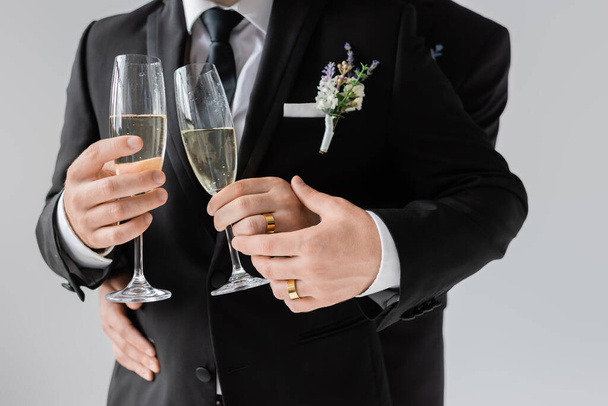 Ausgeschnittene Ansicht eines homosexuellen Paares in klassischen Anzügen und goldenen Ringen, das Champagnergläser hält, während es seine Hochzeit isoliert auf grau feiert  - Foto, Bild