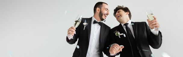 グレーの背景に結婚式のお祝いの間に落ちコンフェッティの下でシャンパンの手と眼鏡を保持している古典的な服装で興奮同性愛者の新郎,バナー  - 写真・画像