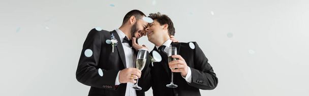 Беззаботные однополые женихи в классах прикасаются друг к другу и держат бокалы шампанского во время празднования свадьбы под падающими праздничными конфетти на сером фоне, баннер  - Фото, изображение