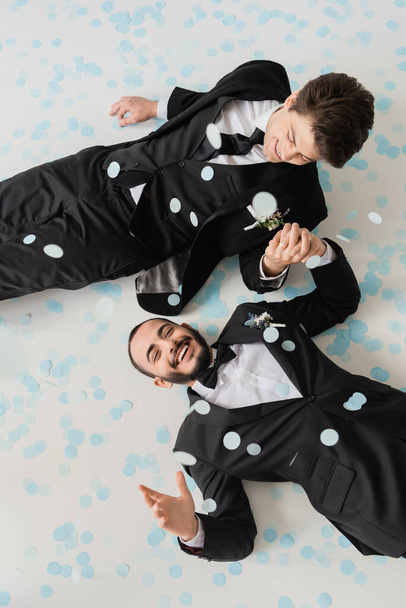 Top view of cheerful same sex couple in classic suits holding hands while having fun and lying on confetti during wedding celebration on grey background  - Photo, Image