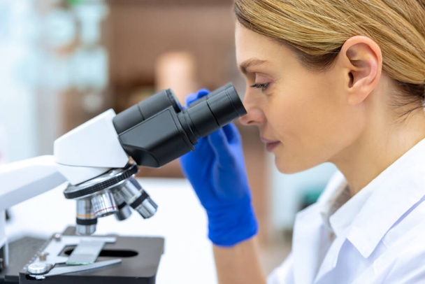 Professional lab. Profile portrait of blonde female medical worker wearing white coat and gloves, using microscope during research. - Foto, Imagen