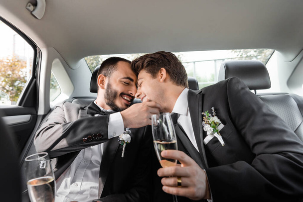 Carefree homosexual groom touching face of young boyfriend in braces and elegant suit with boutonniere and holding champagne while sitting on backseat of car - Foto, afbeelding