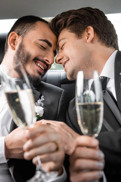Portrait of overjoyed gay newlyweds in classic attire holding blurred hands and glasses of champagne during honeymoon road trip in car  - Photo, Image